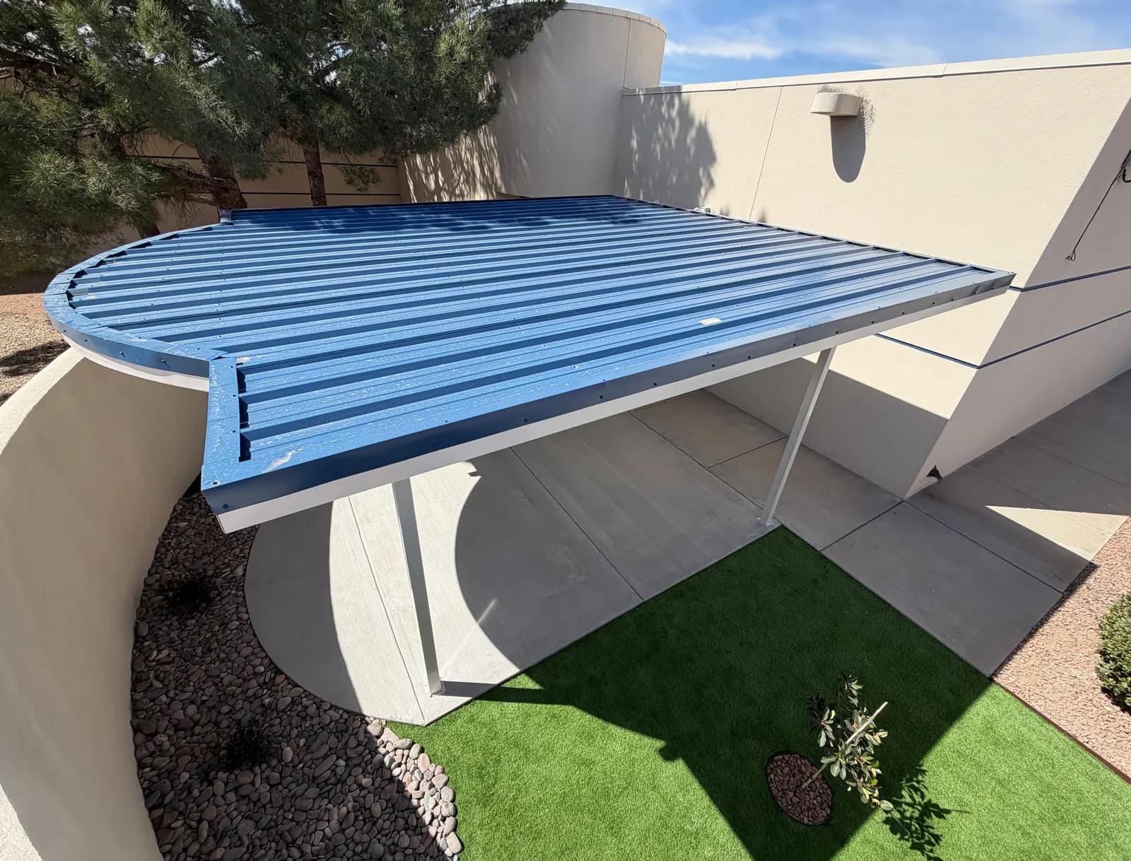 Curved steel shade canopy with blue standing-seam roof over a commercial courtyard in Las Cruces
